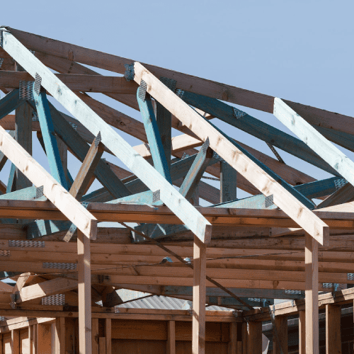 A close-up view of the wooden structural framing for a building's roof, showing pre-fabricated timber roof trusses joined together with galvanized metal connector plates. The unfinished wooden framework stands out against a pale blue sky.