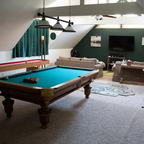 A spacious, carpeted attic game room featuring vaulted ceilings with exposed white support beams. In the foreground, a classic wooden pool table with vibrant teal felt sits lit under three pendant lamps. In the background, a cozy living area includes light brown sofas, a wall-mounted TV against a dark green accent wall, and a shuffleboard table off to the side.