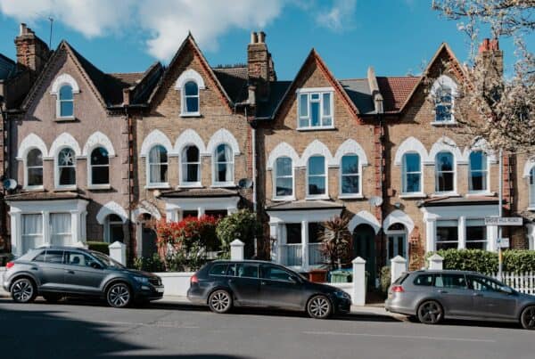 A row of two-story brick Victorian terraced houses featuring steep gables, decorative white trim around pointed arched windows, and bay windows. Cars are parked along the street in front of white garden walls under a bright blue sky on Grove Hill Road.