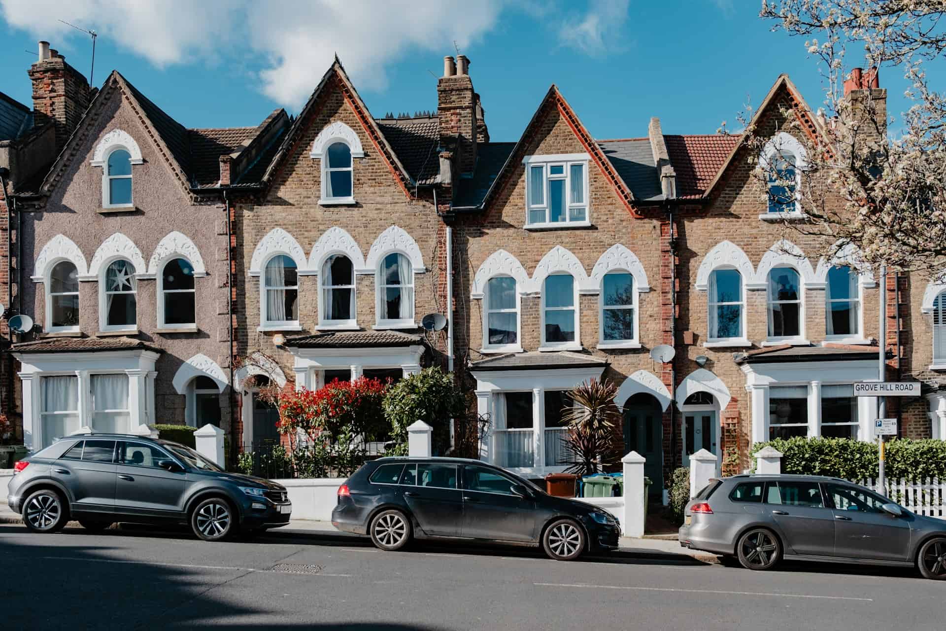 A row of two-story brick Victorian terraced houses featuring steep gables, decorative white trim around pointed arched windows, and bay windows. Cars are parked along the street in front of white garden walls under a bright blue sky on Grove Hill Road.